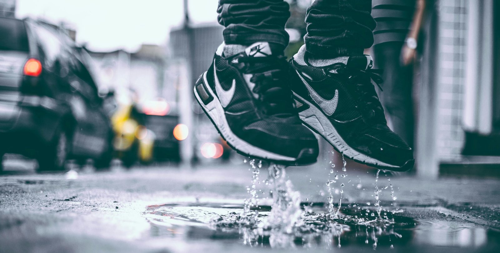 Close-up of a pair of black Nike sneakers splashing in a puddle as the person wearing them jumps in the air, with blurred city background and water droplets frozen mid-air.